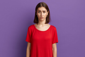 Obraz premium I can not believe. Surprised pretty brunette girl in red t-shirt notices something unexpected, hears bad news. Looks with horror at the camera. Posing over purple background.