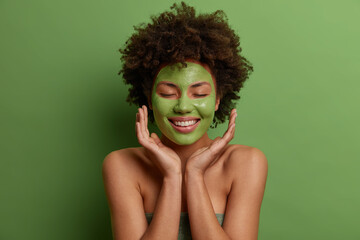 Portrait of curly haired woman smiles gently applies beauty mask keeps hands near face enjoys skin care treatments stands bare shoulders isolated over bright green background. Wellness concept