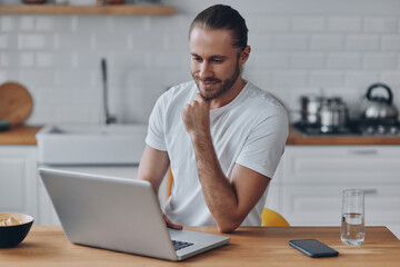 Handsome young man using laptop while working at the kitchen table at home