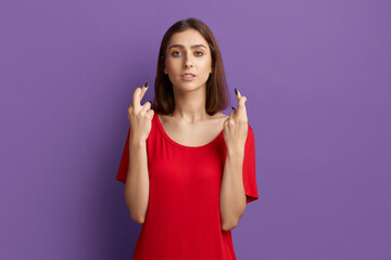 I wish I will win. Excited young pretty brunette girl in red t-shirt crossing fingers in hope gesture. Believe in her lucky day and all dreams come true. Posing over purple background.