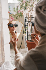 Attractive young woman in bathrobe using lipstick while sitting on the balcony