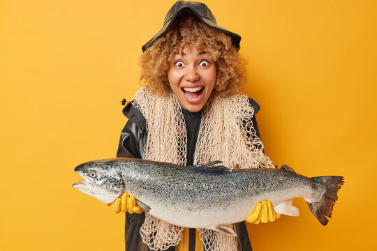 Indoor shot of surprised cheerful curly woman caught very big fish holds large salmon carries fishing net wears black leather raincoat and hat isolated over yellow background. Hobby concept.