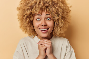 Portrait of surprised cheerful woman keeps hands together smiles toothily reacts to amazing news hears something excellent dressed in casual t shirt isolated over beige background. People emotions