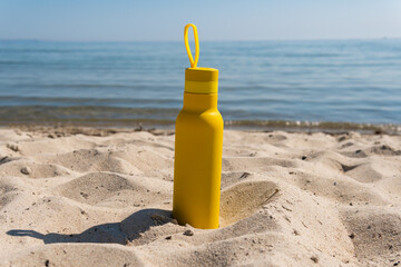 A yellow bottle, thermos standing in the sand on a beach, ocean on sunny summer day. Symbol of summer, holidays and rest
