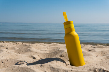 A yellow bottle, thermos standing in the sand on a beach, ocean on sunny summer day. Symbol of...