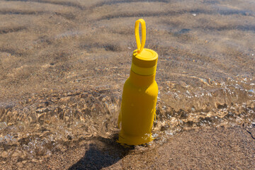 A yellow bottle, thermos standing in the sand on a beach, ocean on sunny summer day. Symbol of summer, holidays and rest