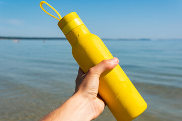 Keeping a yellow bottle in hands, thermos on a beach, ocean on sunny summer day. Symbol of summer, holidays and rest
