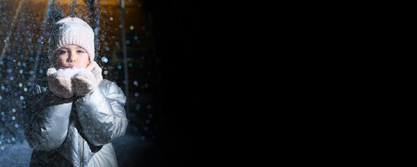 Girl and snow. Girl blowing on palms with snow at night with blue backlight and blurry lights on a black isolated background. Banner