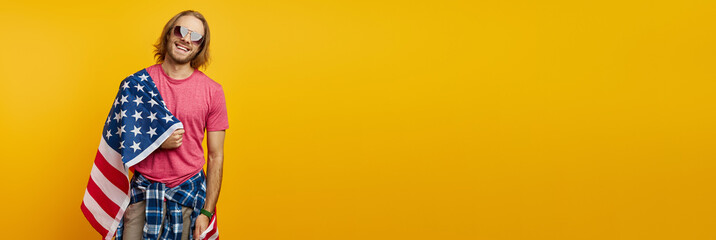 Handsome young man carrying American flag on shoulder and smiling against yellow background