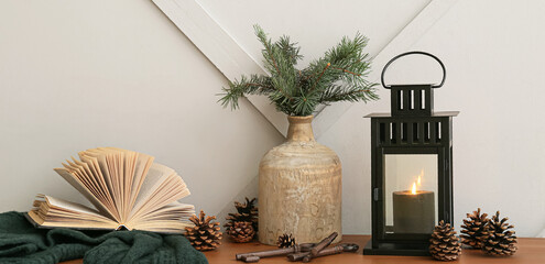 Christmas lantern with burning candle, vase, cones and book on table near light wall © Pixel-Shot
