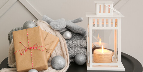 Christmas lantern with burning candle, gift boxes, decor and warm clothes on table near light wall