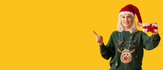 Mature woman with Christmas gift box pointing at something on yellow background with space for text