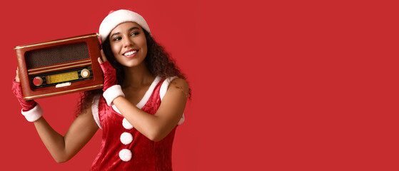 Young African-American woman in Santa costume and with retro radio receiver listening to music on red background with space for text
