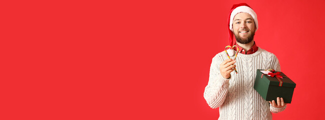 Handsome man with Christmas gift and candy canes on red background with space for text