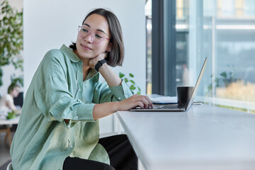 Thoughtful Asian female freelancer works on remote project browses information in web site drinks coffee poses at windowsill in cozy cafe looks away with pensive expression. Professional copywriter
