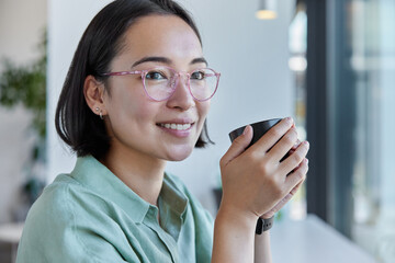 Indoor shot of pretty young Asian woman with dark hair has cheerful satisfied dreamy expression holds mug of tea or coffee looks away happily wears spectacles and shirt being deep in thoughts.