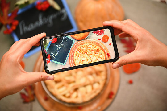 Selective focus. Woman hands hold a smartphone, takes a photo of a freshly baked pumpkin apple pie with a crispy lattice for a food blog. Thanksgiving concept. Autumn holiday. Phone in live view mode