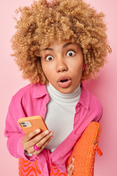 Image of pretty curly woman keeps eyes widely opened jaw dropped holds smartphone reacts to shocking news dressed in casual clothes isolated over pink background. People and human reactions concept