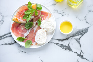 Plate with prosciutto ham, cantaloupe, mozzarella and green basil, top view on a white marble...