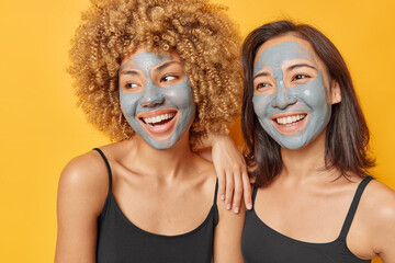 Indoor shot of optimistic mixed race women apply facial clay masks to reduce pores blackheads smile broadly look aside dressed in black t shirts have cheerful expressions isolated on yellow background