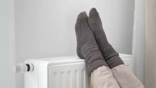 The woman put her frozen feet in woolen warm socks on top of the warm radiator of the home heater. Limiting energy consumption during the cold winter heating period during the global energy crisis.
