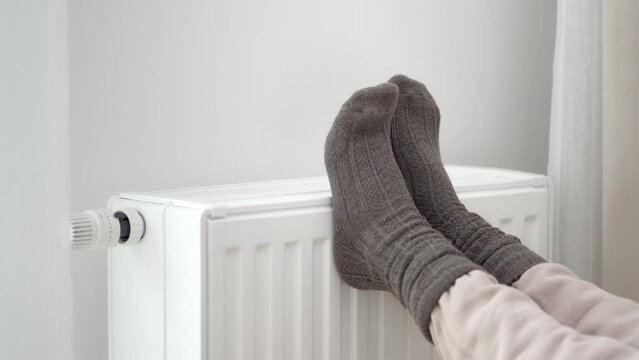 The woman put her frozen feet in woolen warm socks on top of the warm radiator of the home heater. Limiting energy consumption during the cold winter heating period during the global energy crisis.