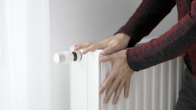 A man touches the cold radiator of a home heater with his frozen hands and increases the heating power by the maximum value. Limiting energy consumption during the heating season. The energy crisis.