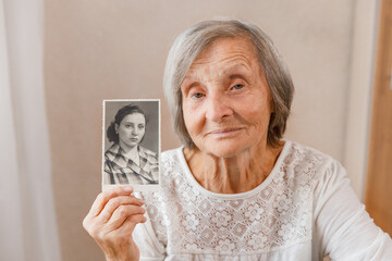 Senior woman is holding a photo of their youthful selves. Memories, nostalgia, time concept The photo was taken in 1958 - obrazy, fototapety, plakaty