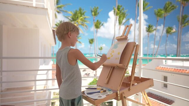 Young blond boy painting at the balcony of a resort hotel room in front of amazing tropical beach with palm trees, turquoise sea water and blue skies