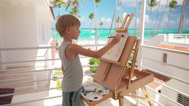 Young blond boy painting at the balcony of a resort hotel room in front of amazing tropical beach with palm trees, turquoise sea water and blue skies