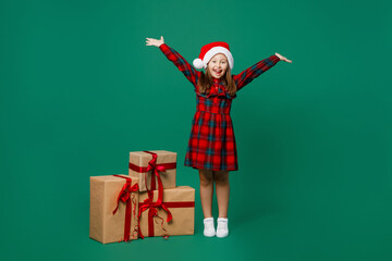 Full body little child kid girl 7 years old wear red dress Christmas hat posing stand near present box gift ribbon bow spread hands isolated on plain dark green background Happy New Year 2023 concept.