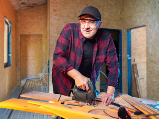 Man is carpenter. Guy with electric jigsaw is engaged in woodworking. Carpenter smiles and looks...