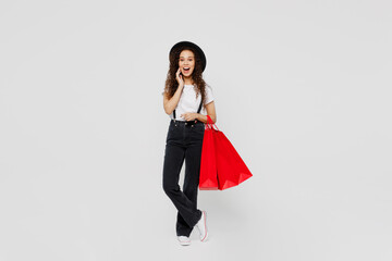 Full body amazed shocked happy young woman wear t-shirt hat hold in hand red paper package bags after shopping touch face isolated on plain solid white background Black Friday sale buy day concept.