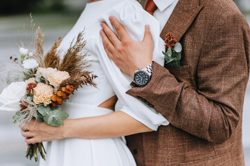 A stylish, fashionable groom in a brown suit gently hugs the bride in a white dress with a bouquet of wildflowers. Wedding photography, close-up portrait.