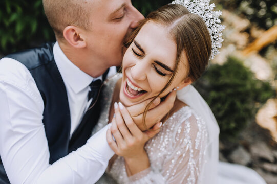 Wedding photo of cheerful, smiling, funny newlyweds. A stylish, young groom and a beautiful bride in a white dress with a bouquet in their hands, a diadem on their heads, sit in a park in nature.
