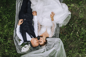 A stylish groom and a beautiful bride with a diadem lie on a plaid with a long white veil, a...