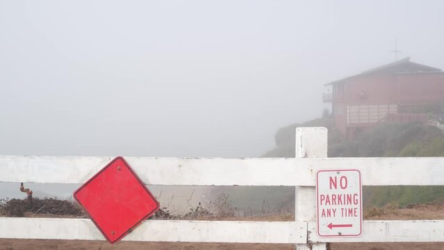 No parking any time road sign in dead end. Road ends by foggy ocean beach, California coast, USA. Misty overcast weather and traffic signage on city street. Red romb on wooden fence by bluff.