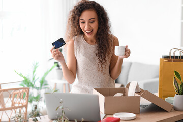 Surprised young African-American woman with credit card and cup of coffee shopping online at home