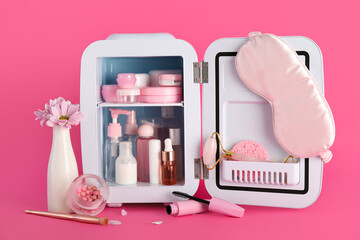 Small refrigerator with cosmetic products and flowers in vase on pink background