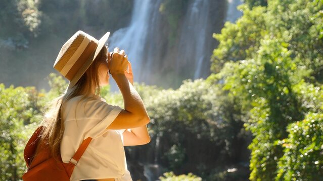 Cinematic shot of happy travel woman on trip or adventure. Young blogger girl millennial traveler make photo for social media of amazing waterfall, nature on trendy retro camera. Wanderlust concept