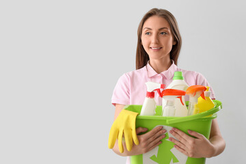 Young woman holding bucket with cleaning supplies on light background