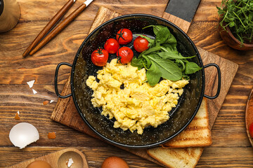 Board with frying pan of scrambled eggs, tomatoes, greens and toasts on wooden background