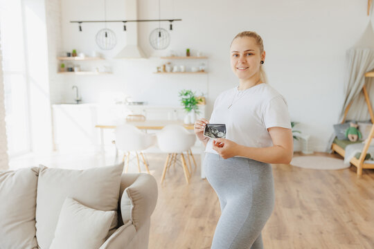 Beautiful blonde pregnancy young woman holding ultrasound scan in front of bright scandinavian kitchen