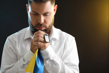 Young man with flag of Ukraine and military tag praying on dark background, closeup