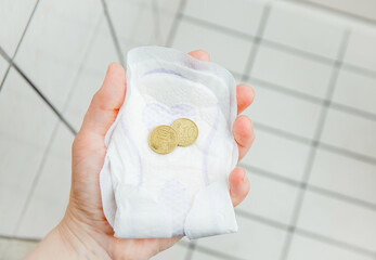 Hygiene poverty concept, young woman holding one time menstrual pad and coins in hand in home bathroom. Can not afford new ones.