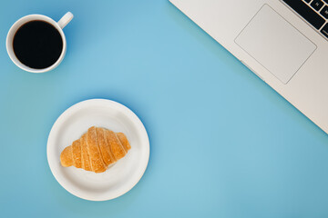 Croissant, cup of coffee and laptop on a blue background, flat lay.
