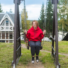 a woman with red hair and a red hoodie is swinging on a wooden swing against the background of a river and tall fir trees.