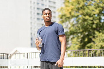 Sportive young thirsty handsome african american man holds plastic bottle of water (isotonic), fresh drink after cardio workout outside, tree and building are on background. Sport. Healthy lifestyle
