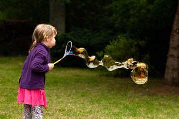 Happy cheerful school age child blowing huge soap bubbles outdoors, young Caucasian girl making a...