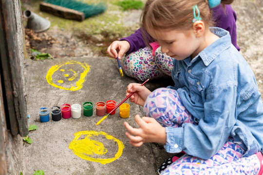 Young children painting on concrete, two girls, art, outdoor activity. Small kids spreading colorful paint on the ground using brushes, outdoors scene, closeup. Child artists, art therapy concept